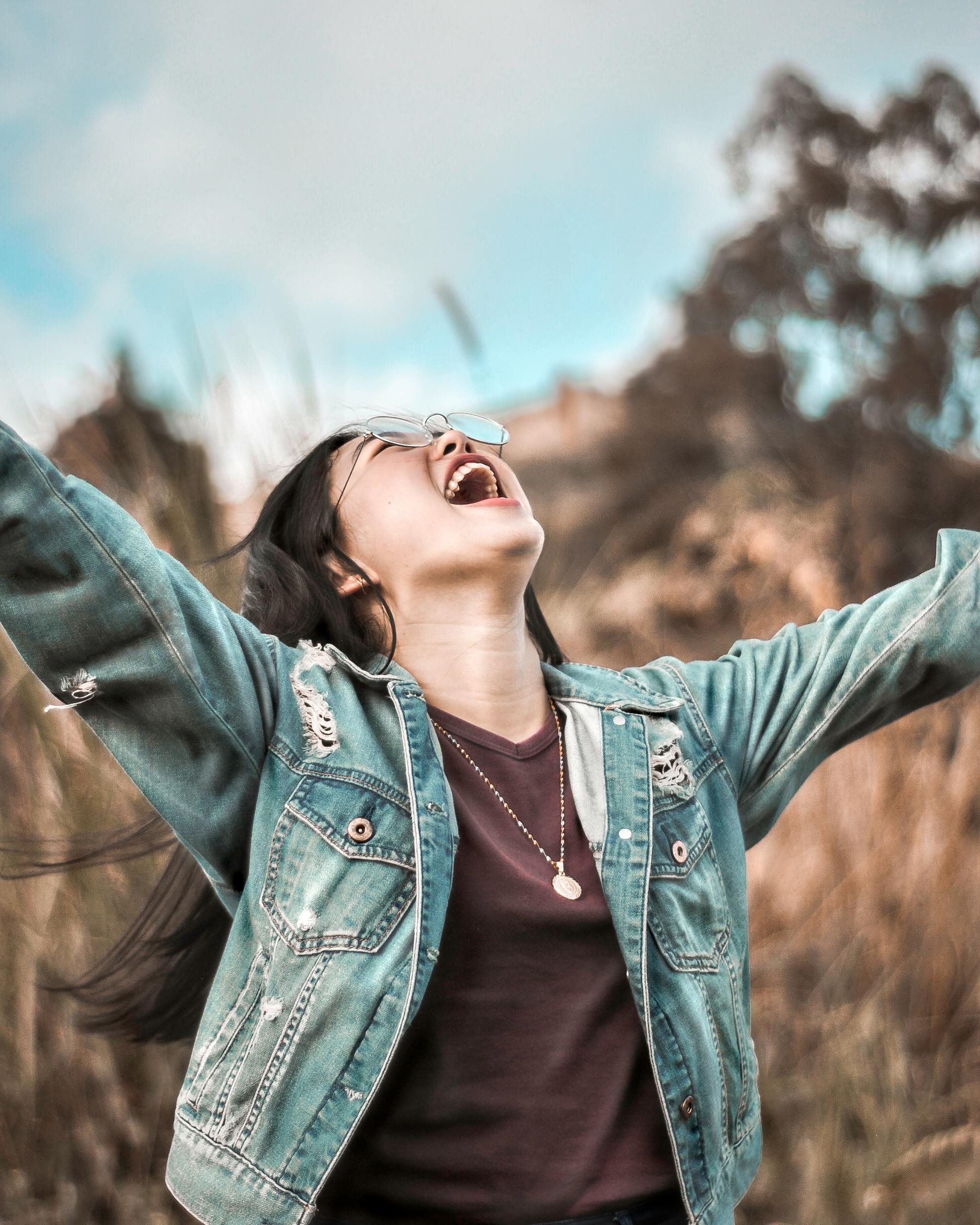 Young woman in denim jacket joyfully celebrating outdoors with arms wide open.