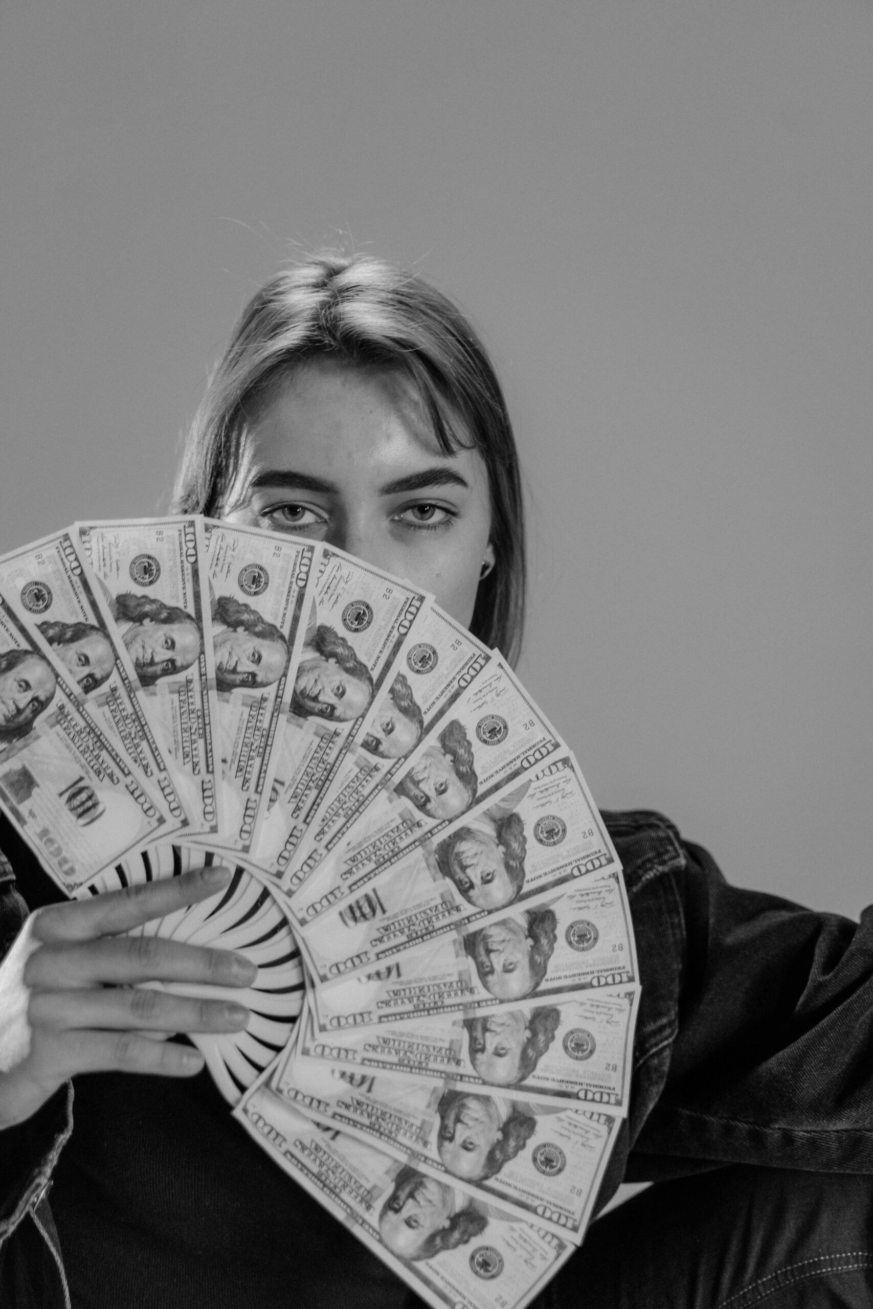 Young woman holding a fan of US dollar bills, partially covering her face, in a black and white portrait.