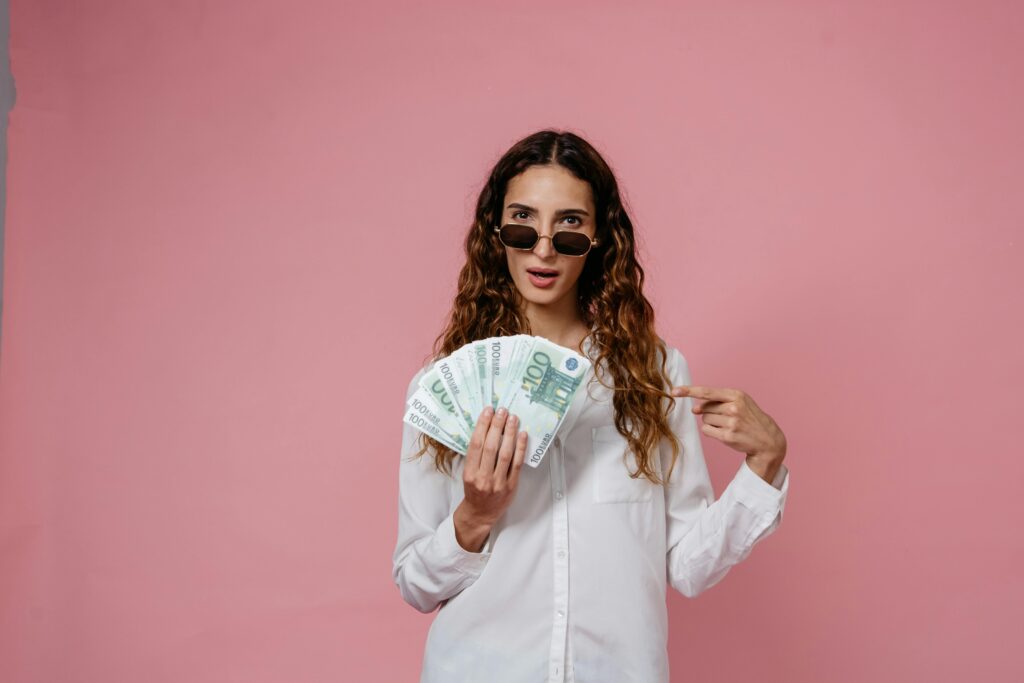 Woman in sunglasses holding and pointing at a stack of 100 euro notes against a pink background.