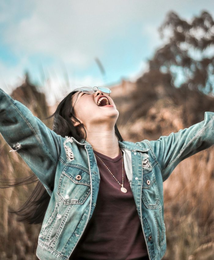 Young woman in denim jacket joyfully celebrating outdoors with arms wide open.