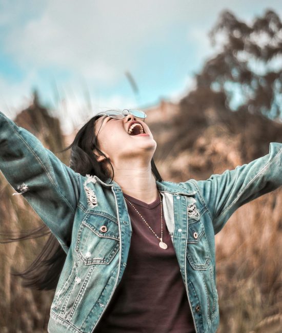 Young woman in denim jacket joyfully celebrating outdoors with arms wide open.