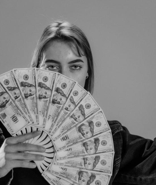 Young woman holding a fan of US dollar bills, partially covering her face, in a black and white portrait.